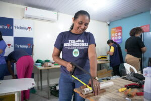 Em uma sala Antônia sorri enquanto utiliza um arco de serra para cortar um cano de PVC branco apoiado sobre uma mesa de madeira. Ela veste uma camiseta azul-marinho com a frase “Terapia Ocupacional” além de calça jeans. Ao fundo, outras mulheres também participam das atividades, lidando com ferramentas e materiais sobre mesas. 