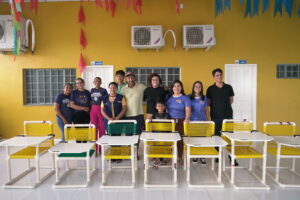 Mariann, presidente do Instituto Pirilampos, posa ao centro de um grupo composto pelos oficineiros responsáveis pela produção das cadeiras. A foto em ambiente interno com decoração de bandeirolas coloridas, paredes amarelas. À frente do grupo, estão as cadeiras produzidas com estrutura de PVC e assentos de corda plástica colorida. O momento marca a entrega oficial dos móveis confeccionados durante a oficina, simbolizando trabalho coletivo, inclusão e compromisso social.