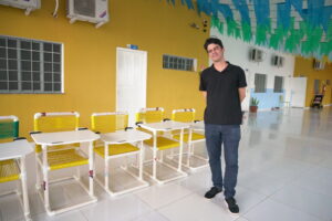 Em um corredor interno de paredes coloridas, Arnaldo aparece de cabelos escuros e curtos posa sorridente ao lado de um conjunto de carteiras escolares infantis feitas com estrutura de PVC e assentos de corda plástica nas cores amarelo e branco. Ele veste uma camisa polo preta, calça jeans escura e tênis pretos com sola branca. O ambiente é bem iluminado, com piso de cerâmica branca brilhante e bandeirolas coloridas (azuis e verdes) penduradas no teto, sugerindo um clima festivo típico de festas juninas.