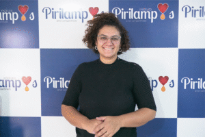 Mariann aparece sorrindo de cabelos cacheados curtos e óculos transparentes, ela está em frente a um painel institucional do Instituto Pirilampos. Ela veste blusa preta de manga curta e brincos circulares. A identidade visual do instituto aparece repetidamente ao fundo, com o slogan "amor compartilhado"